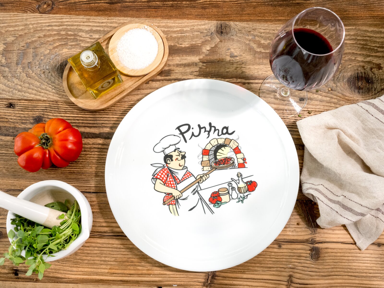 Draufsicht auf einen weißen Pizzateller mit „Pizza“-Schriftzug und Illustration eines Pizzabäckers am Steinofen auf rustikalem Holztisch, umgeben von Tomaten, Kräutern im Mörser, Olivenöl, Salzschale, Rotweinglas und Küchentuch.