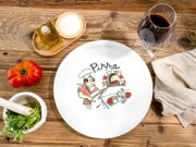 Draufsicht auf einen weißen Pizzateller mit „Pizza“-Schriftzug und Illustration eines Pizzabäckers am Steinofen auf rustikalem Holztisch, umgeben von Tomaten, Kräutern im Mörser, Olivenöl, Salzschale, Rotweinglas und Küchentuch.