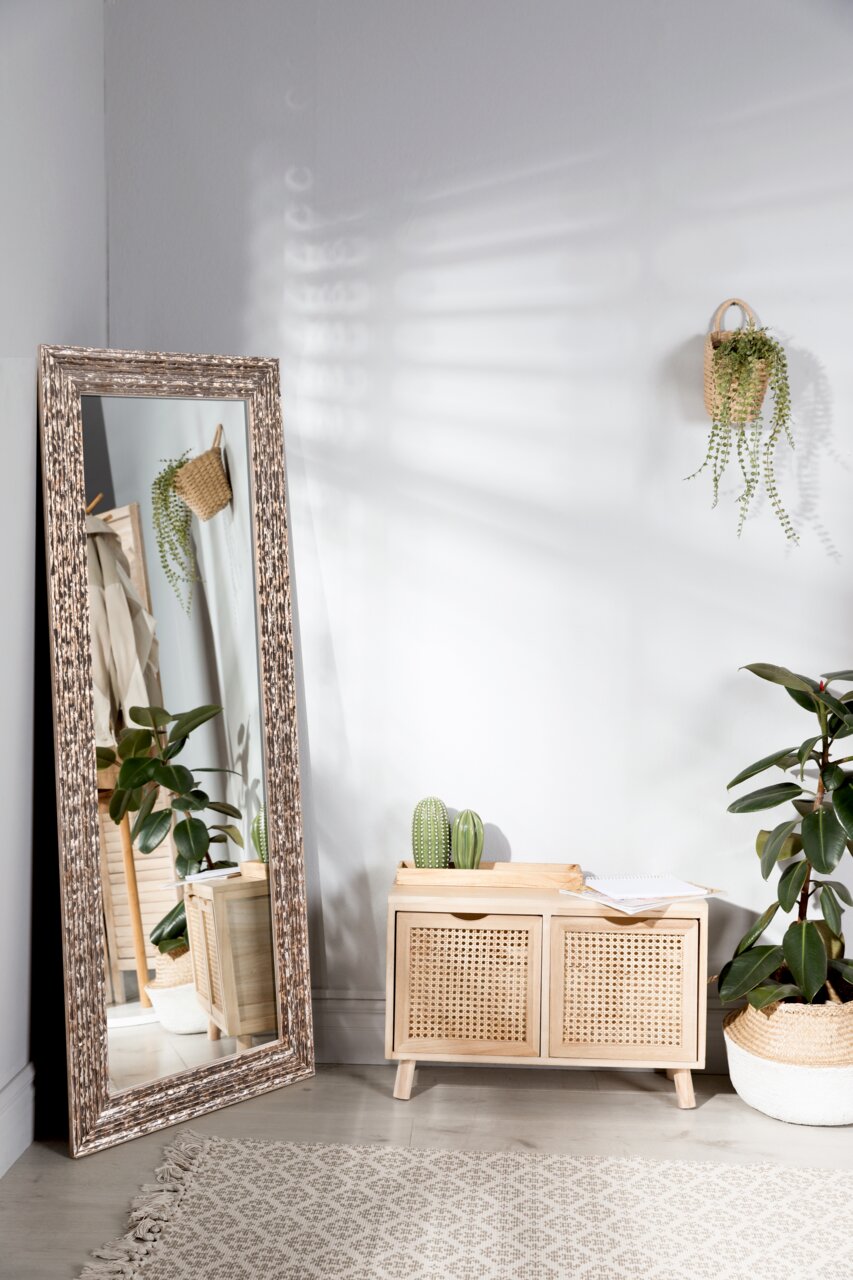 Großer rechteckiger Spiegel mit dekorativem Rahmen, der schräg an einer Wand lehnt. Im Spiegel spiegelt sich ein Teil des Raumes mit Pflanzen und einem kleinen Schrank wider.