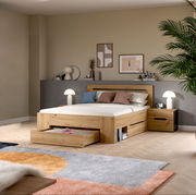 Modernes Holzbett in Eiche-Optik mit gepolstertem Kopfteil, ausziehbarer Bettkasten-Schublade und passendem Nachttisch im Schlafzimmer, fotografiert in frontaler leicht seitlicher Perspektive.
