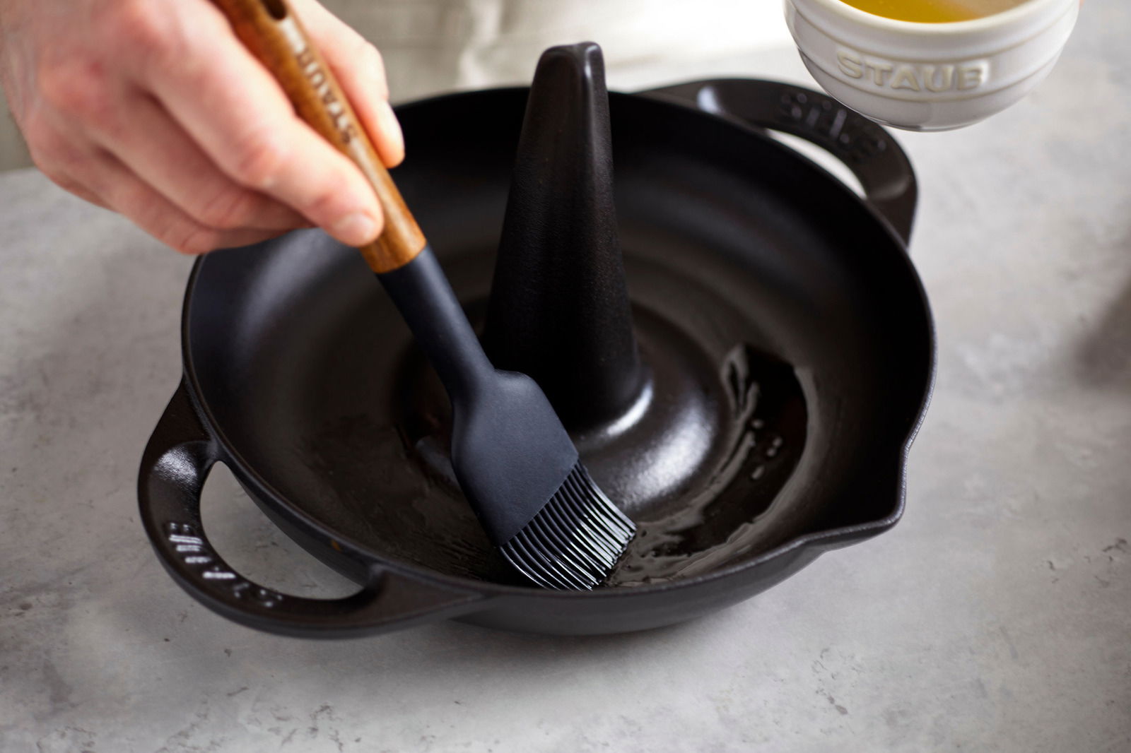 Nahaufnahme eines STAUB Hähnchenbräters mit einem Pinsel, der Öl aufträgt. Der Bräter hat einen zentralen Kegel und zwei Griffe.