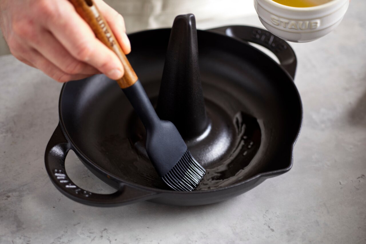Close-up van een STAUB-kippenbraadpan met een kwast waarmee olie wordt aangebracht. De braadpan heeft een centrale kegel en twee handvatten.