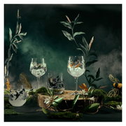 Ein Set aus zwei Gingläsern mit kunstvollen Naturmotiven, umgeben von dekorativen Pflanzen und Nebel, aus einer frontalen Perspektive fotografiert.