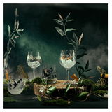 Ein Set aus zwei Gingläsern mit kunstvollen Naturmotiven, umgeben von dekorativen Pflanzen und Nebel, aus einer frontalen Perspektive fotografiert.