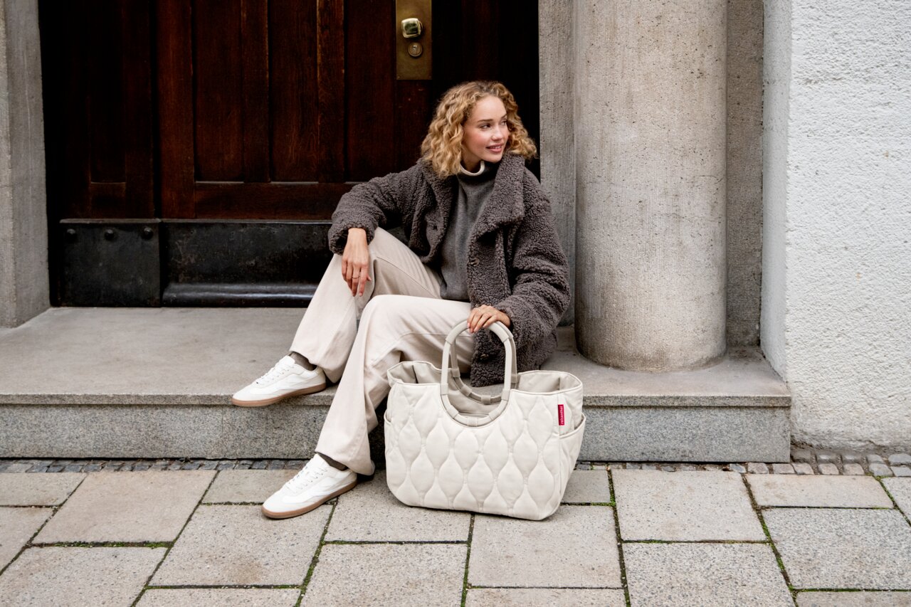 Lifestylefoto in licht zijaanzicht: een vrouw zit op een stenen trede voor een houten deur naast haar staat een crèmekleurige, gewatteerde tas (doodskist-/boodschappentas) met korte hengsels.
