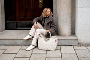 Lifestylefoto in licht zijaanzicht: een vrouw zit op een stenen trede voor een houten deur; naast haar staat een crèmekleurige, gewatteerde tas (doodskist-/boodschappentas) met korte hengsels.