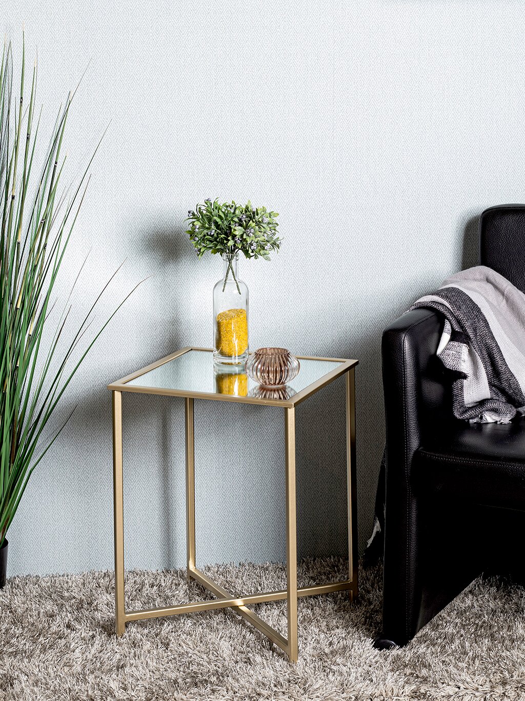 Quadratischer Beistelltisch mit goldfarbenem Metallgestell und Glasplatte, seitliche Perspektive im Wohnzimmer neben schwarzem Sofa; darauf Vase mit grünen Zweigen und kleines Glas-Windlicht.