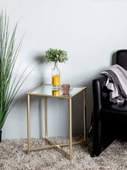 Quadratischer Beistelltisch mit goldfarbenem Metallgestell und Glasplatte, seitliche Perspektive im Wohnzimmer neben schwarzem Sofa; darauf Vase mit grünen Zweigen und kleines Glas-Windlicht.