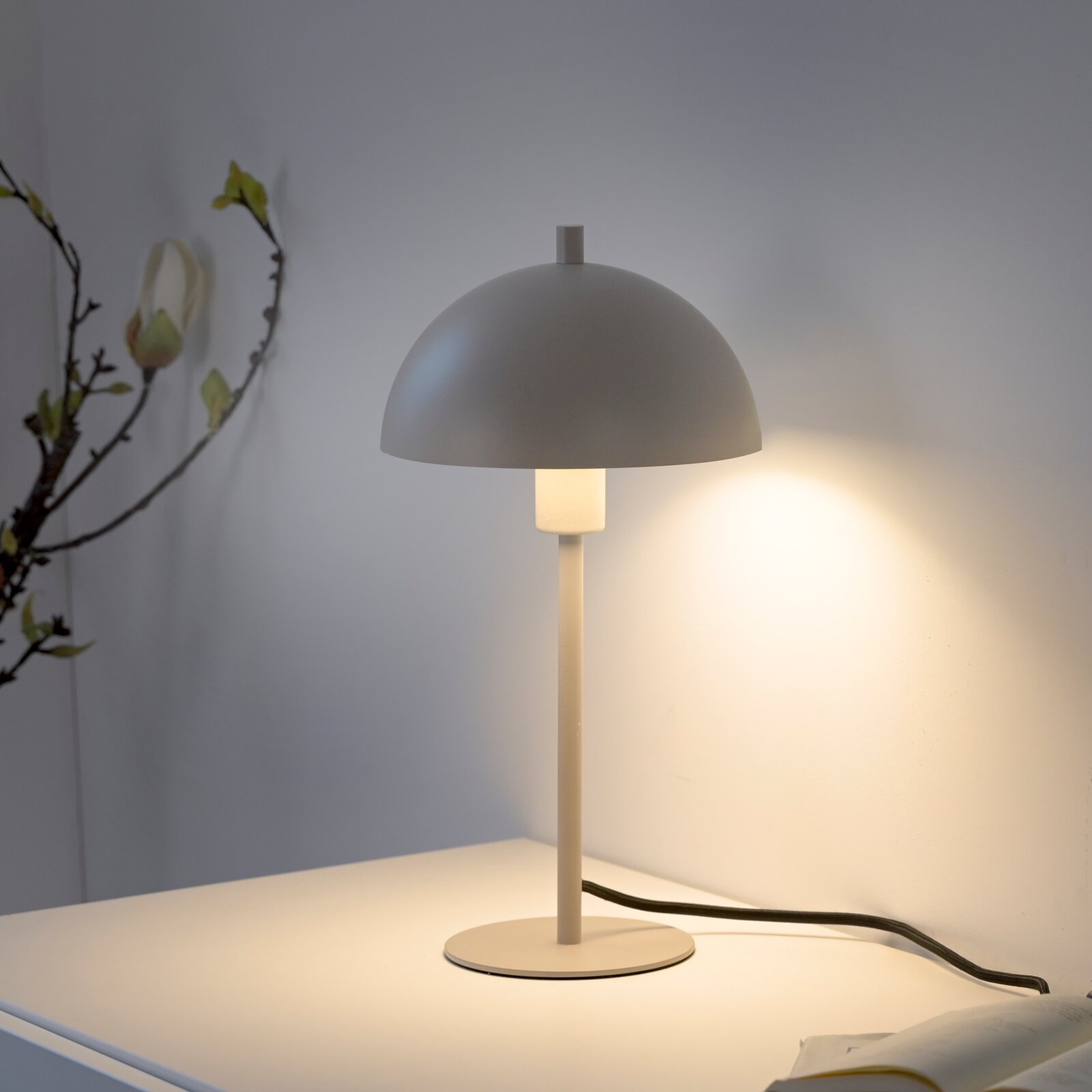 Minimalistische Tischlampe in Beige mit halbkugelförmigem Schirm und warmem Licht auf weißem Tisch, seitliche Frontansicht im leicht schrägen Winkel
