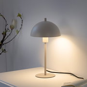 Minimalistische Tischlampe in Beige mit halbkugelförmigem Schirm und warmem Licht auf weißem Tisch, seitliche Frontansicht im leicht schrägen Winkel