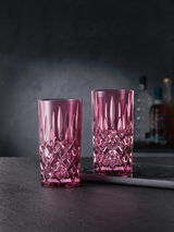 Zwei elegante Longdrinkgläser im Noblesse-Design, in einem kräftigen Pinkton, auf einer dunklen Oberfläche stehend, mit einer stilvollen, facettierten Struktur. Seitliche Perspektive.
