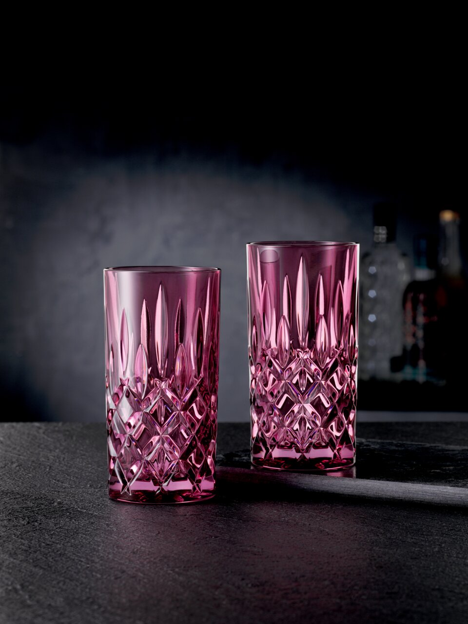Nachtmann Longdrinkglas Noblesse 2er-Set NOBLESSE Zwei elegante Longdrinkgläser im Noblesse-Design, in einem kräftigen Pinkton, auf einer dunklen Oberfläche stehend, mit einer stilvollen, facettierten Struktur. Seitliche Perspektive.