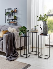 Wohnzimmer-Setting mit drei schwarzen Metall-Pflanzsäulen/Beistelltischen in unterschiedlichen Höhen neben einem grauen Sofa, darauf Deko, Bücher und eine große gemusterte Vase; Perspektive: leicht schräg von vorn auf Augenhöhe.