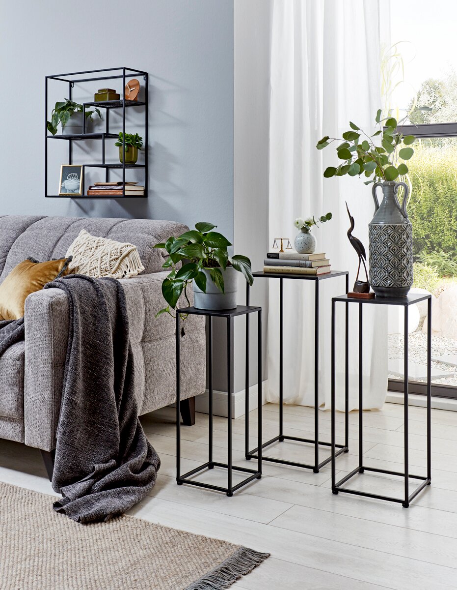 Wohnzimmer-Setting mit drei schwarzen Metall-Pflanzsäulen/Beistelltischen in unterschiedlichen Höhen neben einem grauen Sofa, darauf Deko, Bücher und eine große gemusterte Vase; Perspektive: leicht schräg von vorn auf Augenhöhe.