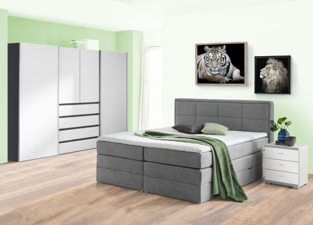 Graues Boxspringbett in einem modernen Schlafzimmer mit grüner Wand, flankiert von einem weißen Nachttisch und einem großen Kleiderschrank. Perspektive von vorne links.