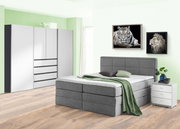 Graues Boxspringbett in einem modernen Schlafzimmer mit grüner Wand, flankiert von einem weißen Nachttisch und einem großen Kleiderschrank. Perspektive von vorne links.