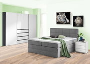 Grijs boxspringbed in een moderne slaapkamer met een groene muur, geflankeerd door een wit nachtkastje en een grote kledingkast. Aanzicht van linksvoor.