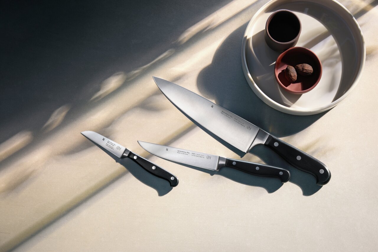 Draufsicht auf ein dreiteiliges Messerset mit schwarzen Griffen, arrangiert neben einer Schale mit zwei kleinen Gefäßen.