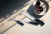 Draufsicht auf ein dreiteiliges Messerset mit schwarzen Griffen, arrangiert neben einer Schale mit zwei kleinen Gefäßen.