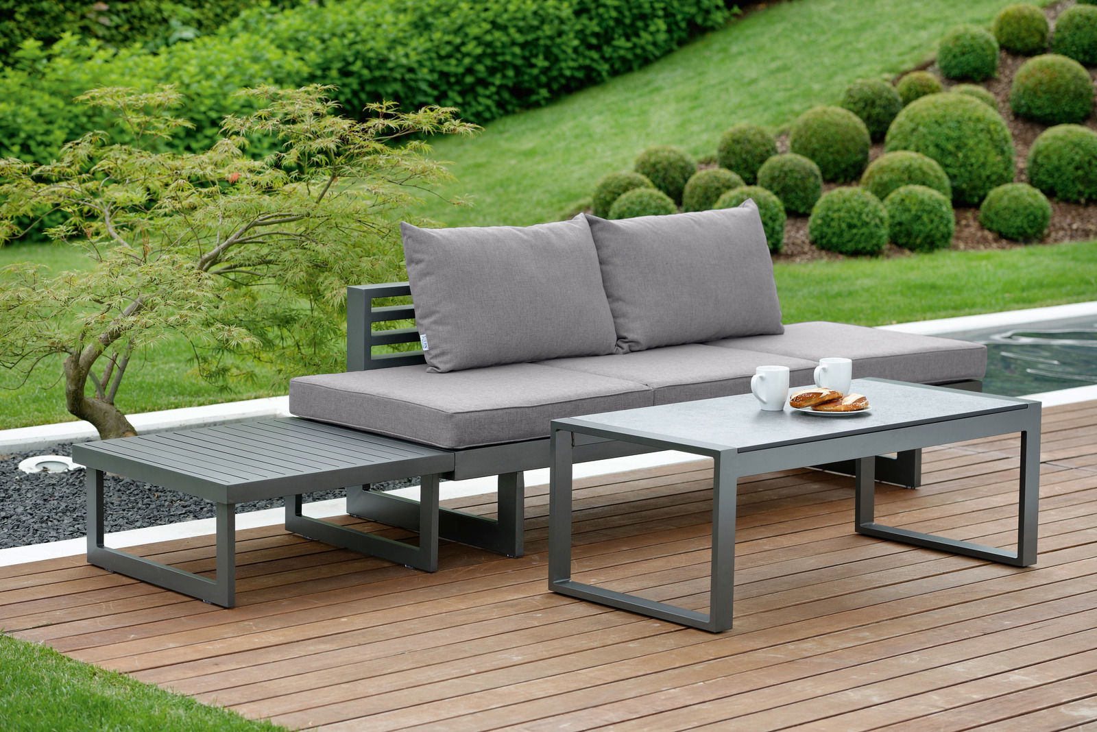Moderne Lounge-Sitzgruppe im Garten mit grauem Sofa, Couchtisch und Beistelltisch, seitliche Perspektive