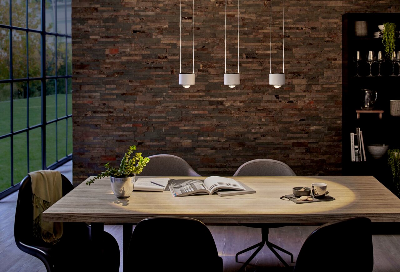 PAULMANN Schienenpendel Aldan URAIL Drei moderne Pendelleuchten über einem Esstisch in einem stilvoll eingerichteten Raum mit Backsteinwand. Perspektive von vorne.
