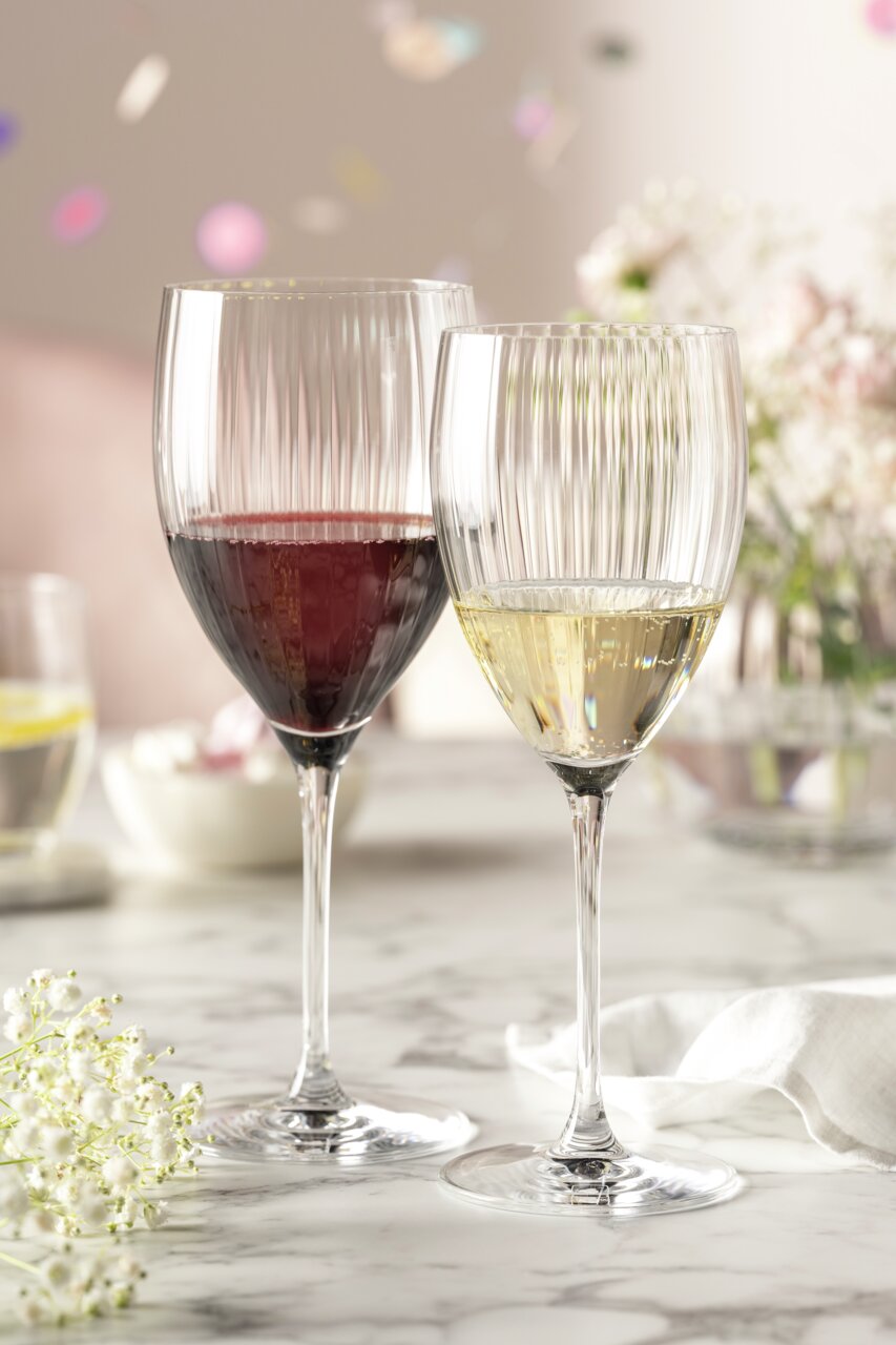 Zwei elegante Weingläser auf einem Marmortisch, eines mit Rotwein und eines mit Weißwein gefüllt, umgeben von zarten Blumen und einem festlichen Ambiente. Seitliche Perspektive.