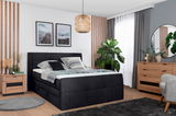 Modernes Schlafzimmer mit einem Boxspringbett in dunklem Stoffbezug, umgeben von stilvollen Holzmöbeln und Pflanzen, aus einer seitlichen Perspektive betrachtet.