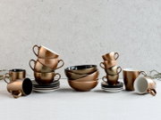 Modernes Keramik-Geschirrset in Metallic-Kupferoptik mit Tassen, Schalen und Untertassen auf grauem Hintergrund, Frontansicht auf Tischhöhe.