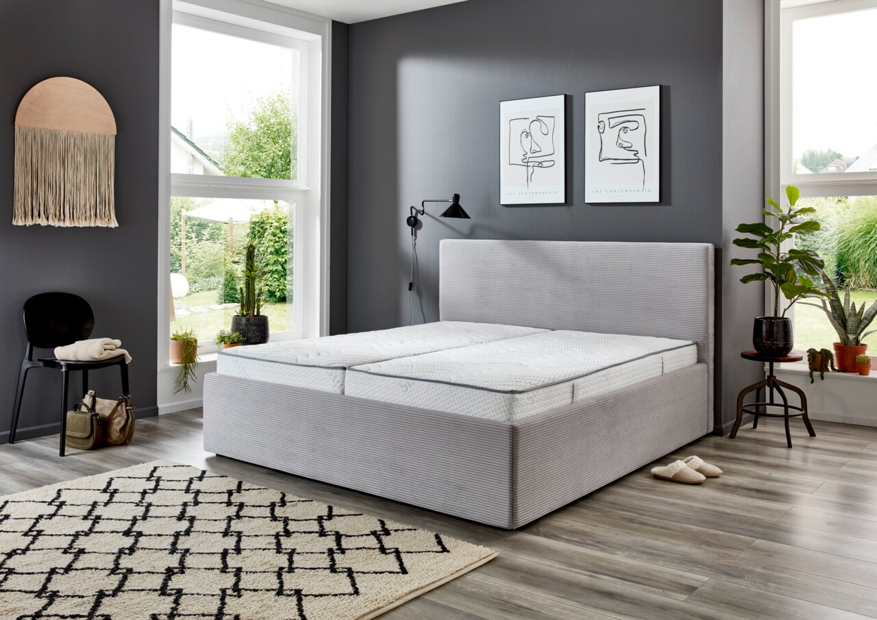 Modernes Polsterbett 180x200 cm in einem stilvoll eingerichteten Schlafzimmer mit grauer Wandfarbe, großen Fenstern und Pflanzen, seitliche Perspektive.