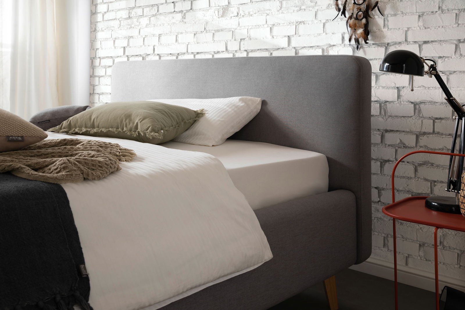 Graues Polsterbett mit Kopfteil, seitliche Perspektive, dekoriert mit Kissen und Decken, neben einem roten Beistelltisch und einer schwarzen Lampe, vor einer weißen Backsteinwand.
