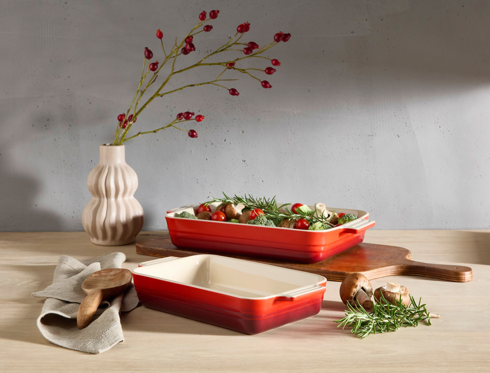 LE CREUSET Auflaufform 32cm schwarz Zwei kirschrote Auflaufformen, eine gefüllt mit frischem Gemüse, auf einem Holztisch. Im Hintergrund eine Vase mit roten Beerenzweigen. Perspektive von vorne.