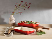 Zwei kirschrote Auflaufformen, eine gefüllt mit frischem Gemüse, auf einem Holztisch. Im Hintergrund eine Vase mit roten Beerenzweigen. Perspektive von vorne.