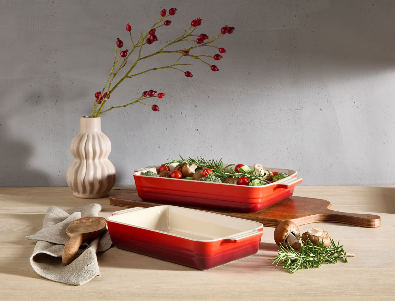 LE CREUSET Auflaufform 32cm schwarz Zwei kirschrote Auflaufformen, eine gefüllt mit frischem Gemüse, auf einem Holztisch. Im Hintergrund eine Vase mit roten Beerenzweigen. Perspektive von vorne.