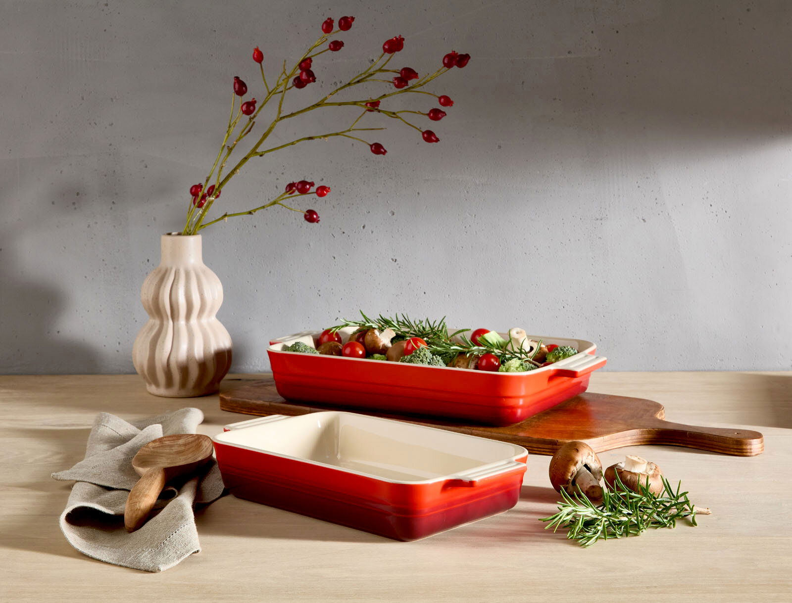 LE CREUSET Auflaufform 32cm schwarz Zwei kirschrote Auflaufformen, eine gefüllt mit frischem Gemüse, auf einem Holztisch. Im Hintergrund eine Vase mit roten Beerenzweigen. Perspektive von vorne.