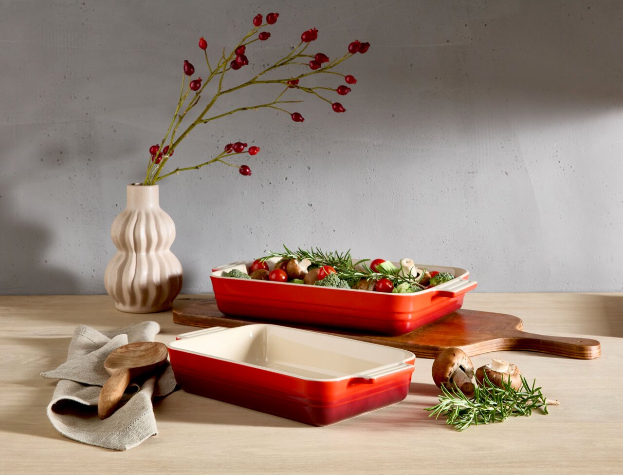 LE CREUSET Auflaufform 32cm schwarz Zwei kirschrote Auflaufformen, eine gefüllt mit frischem Gemüse, auf einem Holztisch. Im Hintergrund eine Vase mit roten Beerenzweigen. Perspektive von vorne.