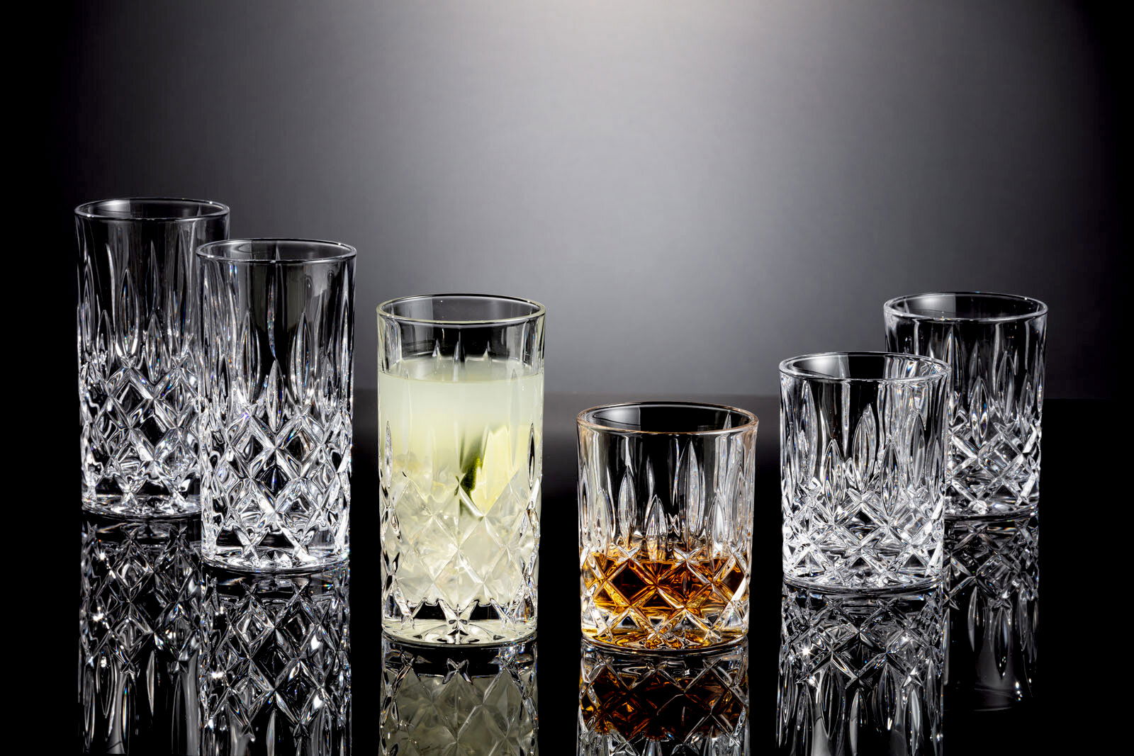 Sechs elegante Kristallgläser der Noblesse-Serie, darunter Whisky- und Longdrinkbecher, auf einer glänzenden Oberfläche mit seitlicher Perspektive.