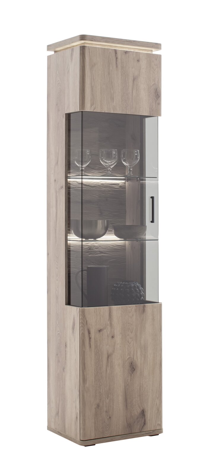 Hoge, moderne vitrinekast in een lichte houtlook met glazen deur, glazen planken en ledverlichting, gevuld met glazen en schalen, gefotografeerd vanuit een licht zijdelings perspectief tegen een witte achtergrond