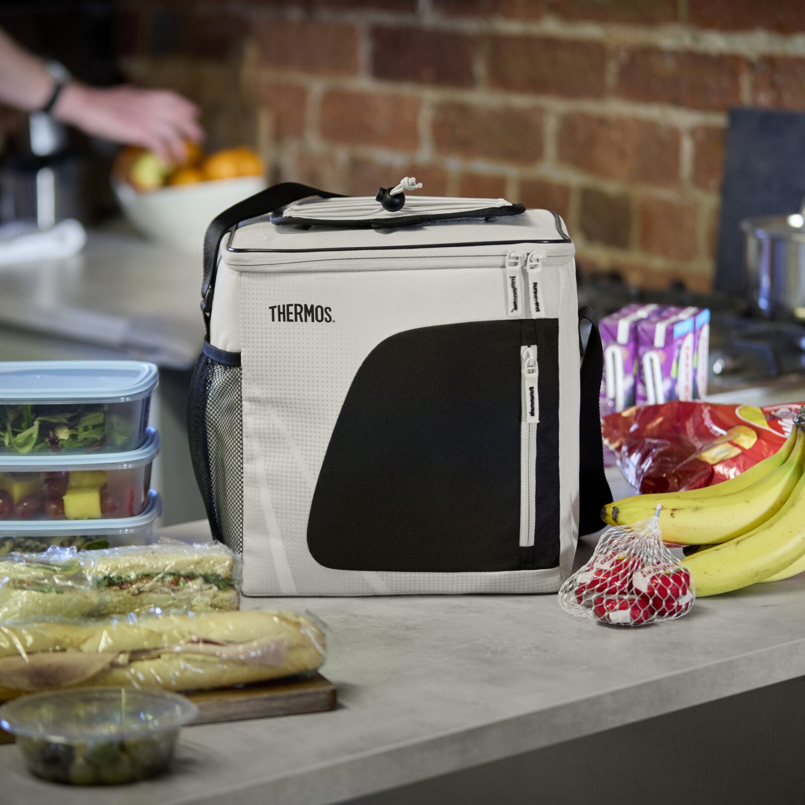 Thermos koeltas in grijs en zwart, vanuit schuin vooraanzicht op een keukenblad tussen lunchboxen, broodjes, fruit en snacks