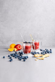 Zwei Kaffeegläser mit rotem Smoothie, dekoriert mit Heidelbeeren, auf einem Tisch mit Früchten und Ingwer, seitliche Perspektive.