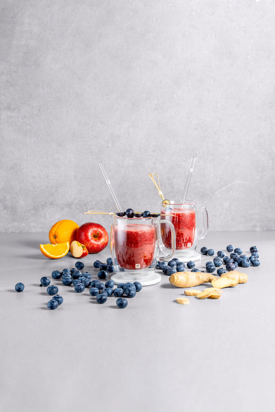 Zwei Kaffeegläser mit rotem Smoothie, dekoriert mit Heidelbeeren, auf einem Tisch mit Früchten und Ingwer, seitliche Perspektive.