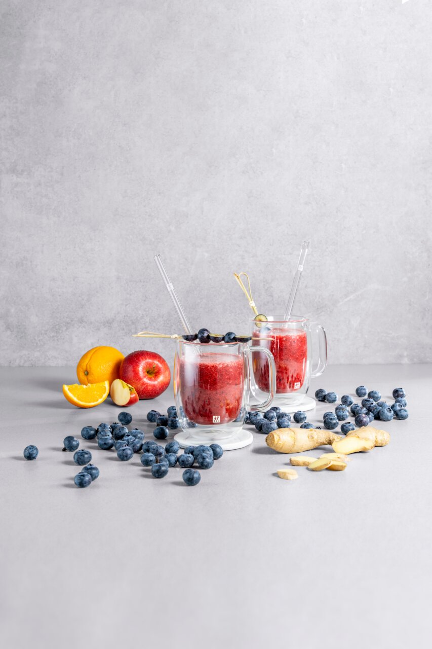 Zwei Kaffeegläser mit rotem Smoothie, dekoriert mit Heidelbeeren, auf einem Tisch mit Früchten und Ingwer, seitliche Perspektive.