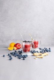 Zwei Kaffeegläser mit rotem Smoothie, dekoriert mit Heidelbeeren, auf einem Tisch mit Früchten und Ingwer, seitliche Perspektive.