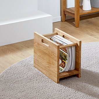 Zurbrüggen Eiche-Massivholz Zeitungsständer Schräg von vorne fotografierter Magazinhalter aus hellem Holz mit seitlichem Griffloch, gefüllt mit Zeitschriften, auf einem hellen Teppich im Wohnraum.