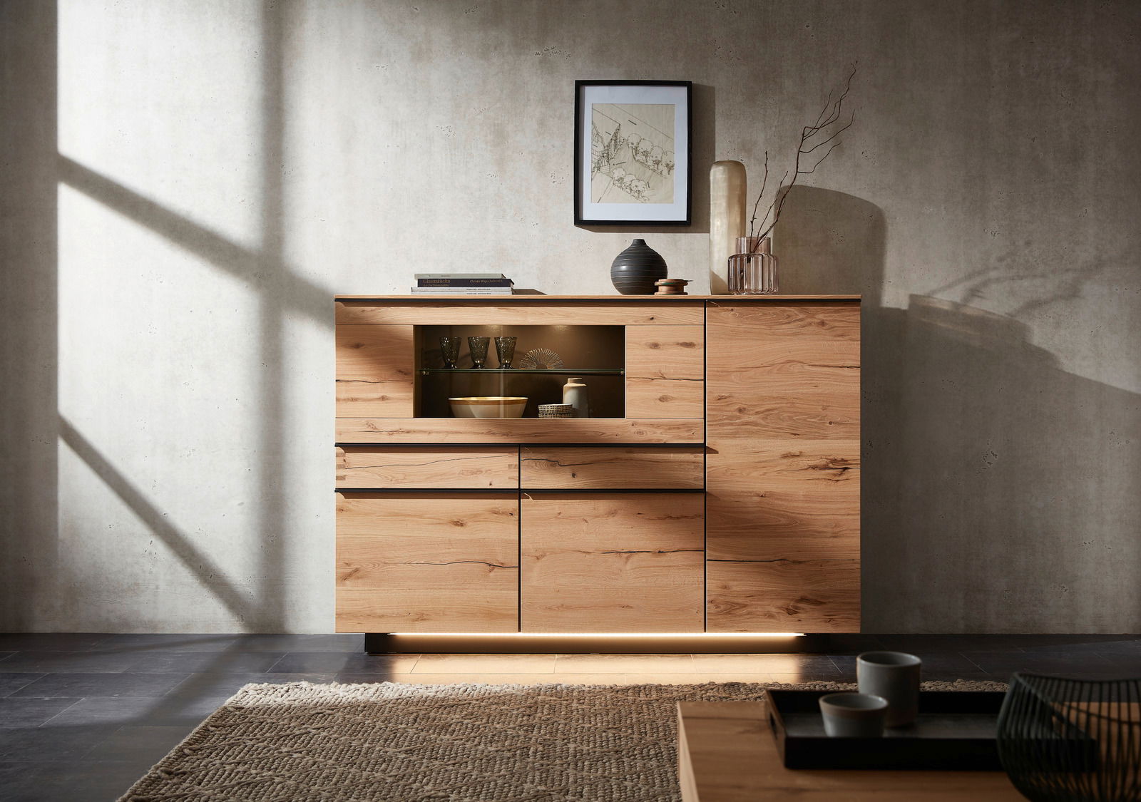Modernes Highboard aus Holz mit dekorativen Elementen, frontal fotografiert. Es hat eine Glasvitrine und ist von natürlichem Licht beleuchtet.