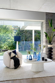 Gemütlicher, beiger Sessel mit Kissen und Decke, frontal betrachtet, in einem modernen Wohnzimmer mit großen Fenstern und Blick auf den Garten.