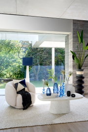 Gemütlicher, beiger Sessel mit Kissen und Decke, frontal betrachtet, in einem modernen Wohnzimmer mit großen Fenstern und Blick auf den Garten.