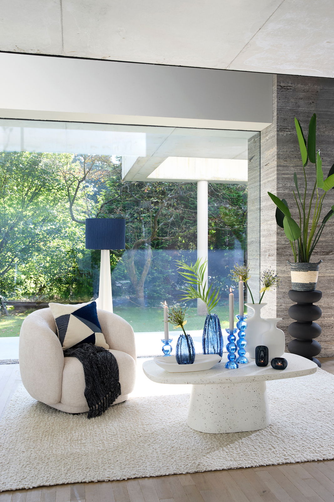 Gemütlicher, beiger Sessel mit Kissen und Decke, frontal betrachtet, in einem modernen Wohnzimmer mit großen Fenstern und Blick auf den Garten.