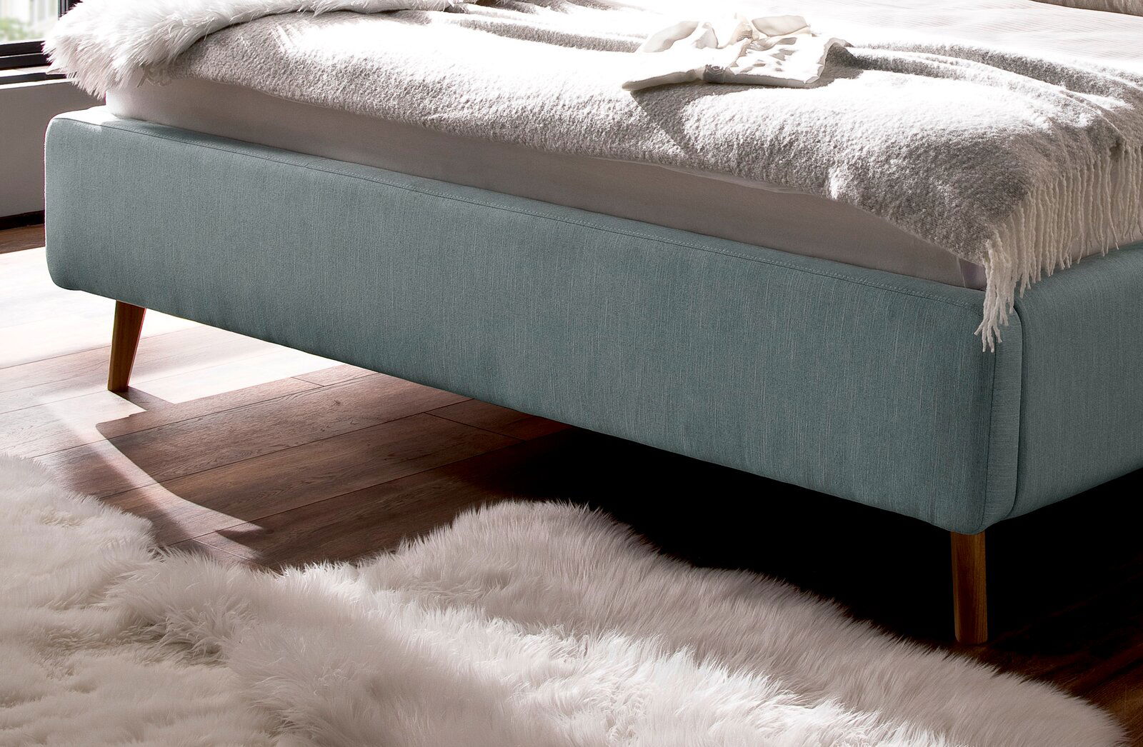 Seitliche Perspektive eines Polsterbetts mit hellblauem Stoffbezug und Holzfüßen auf einem Holzboden mit flauschigem Teppich.