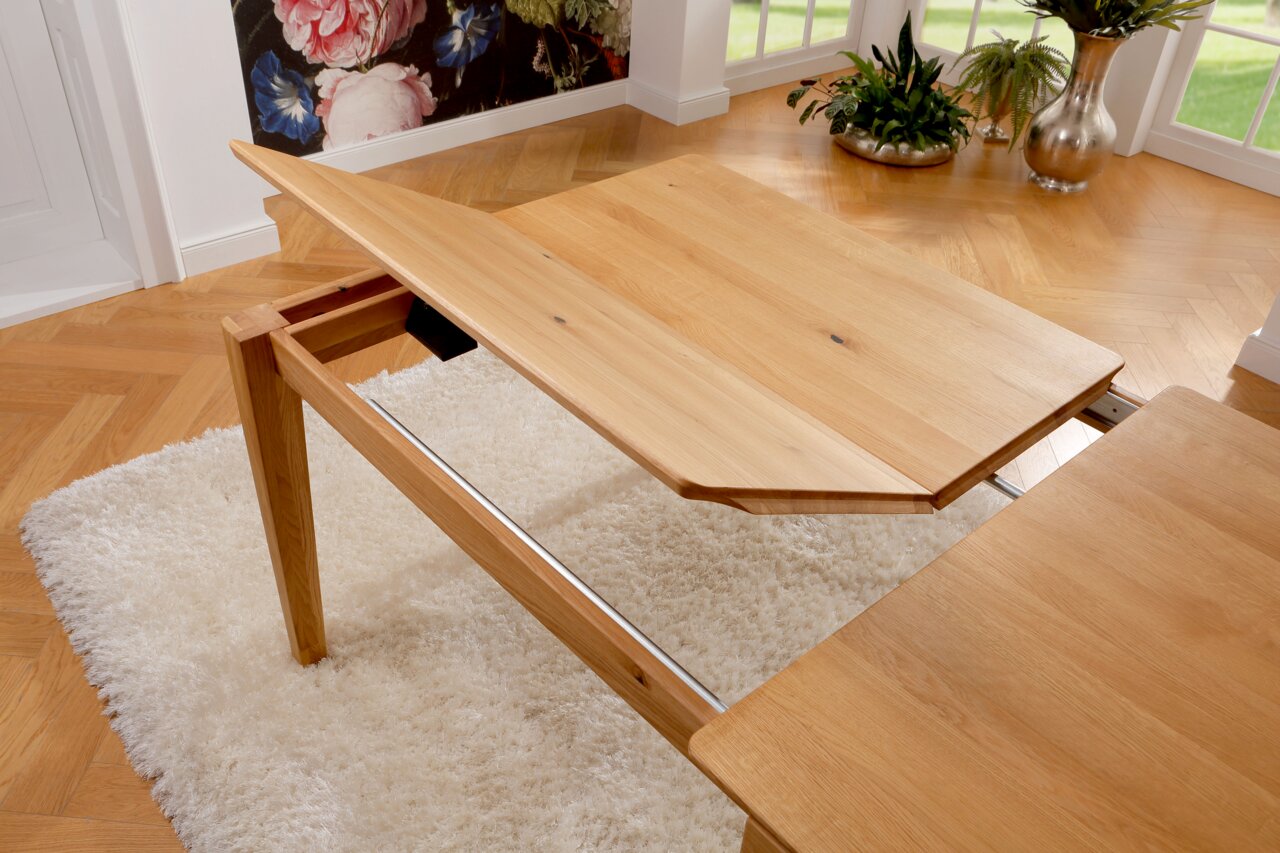 Esstisch aus Holz mit ausziehbarer Platte, seitliche Perspektive, auf einem weißen Teppich in einem hellen Raum mit Pflanzen.