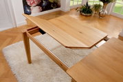 Esstisch aus Holz mit ausziehbarer Platte, seitliche Perspektive, auf einem weißen Teppich in einem hellen Raum mit Pflanzen.